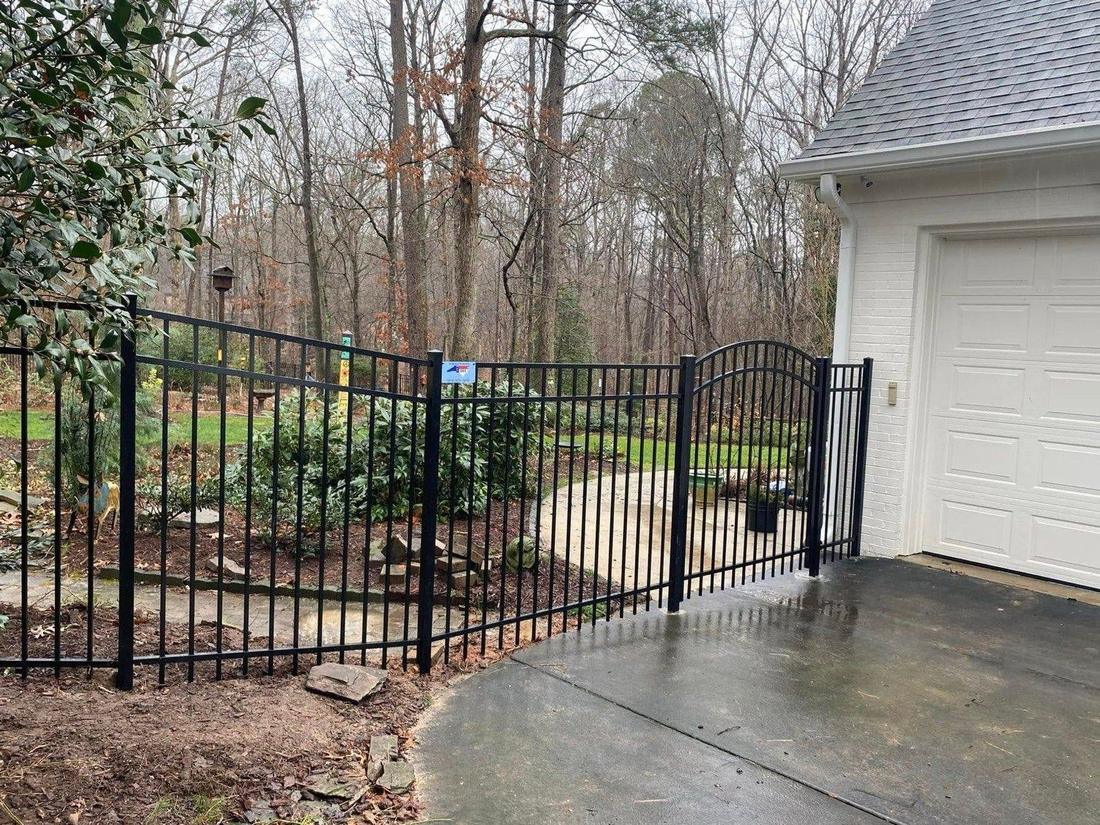 A black aluminum fence with an arched-top gate beside a garage, on a wooded lot after rain.