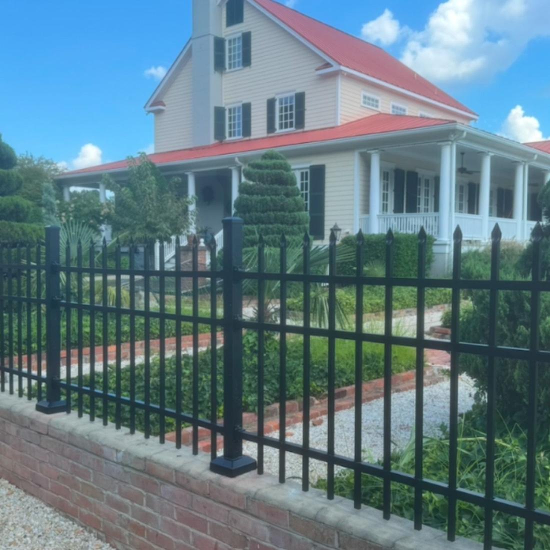 A black aluminum fence set on a brick wall, with a white house and red metal roof behind.