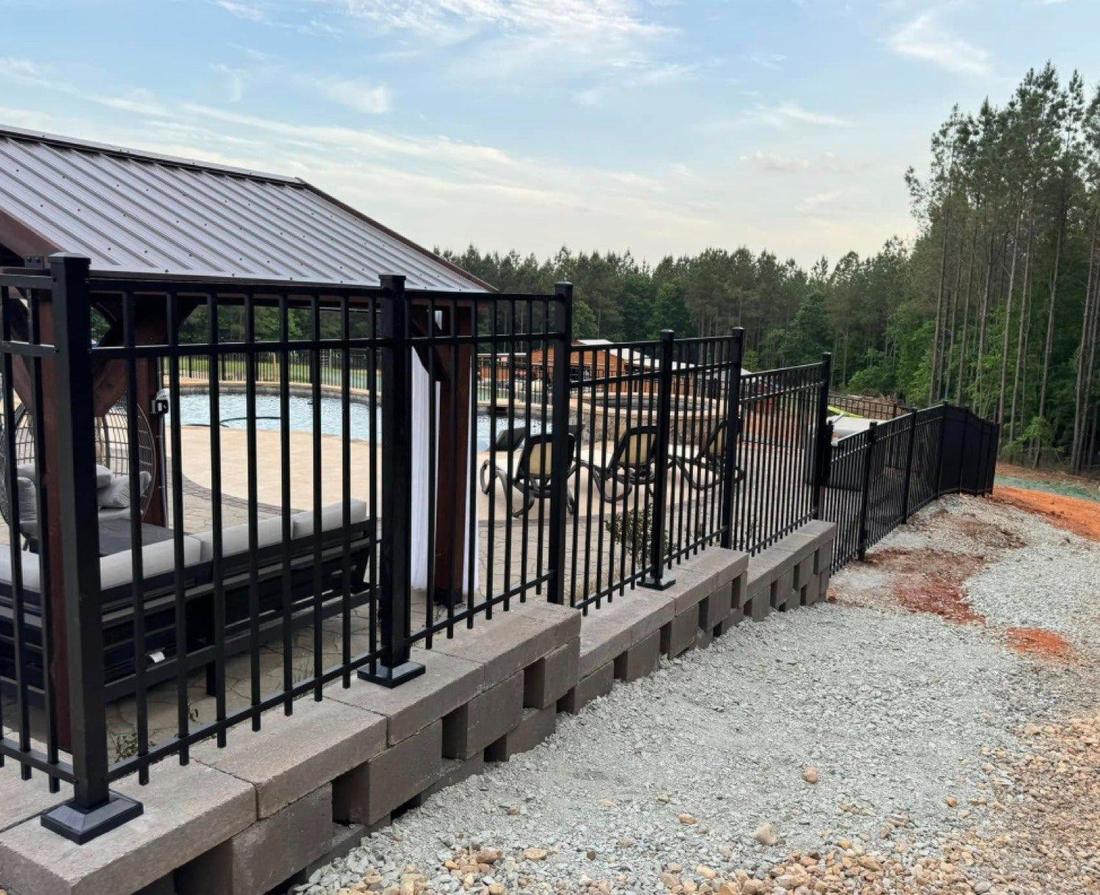 A black aluminum fence on a block retaining wall enclosing a pool and a covered pavilion.
