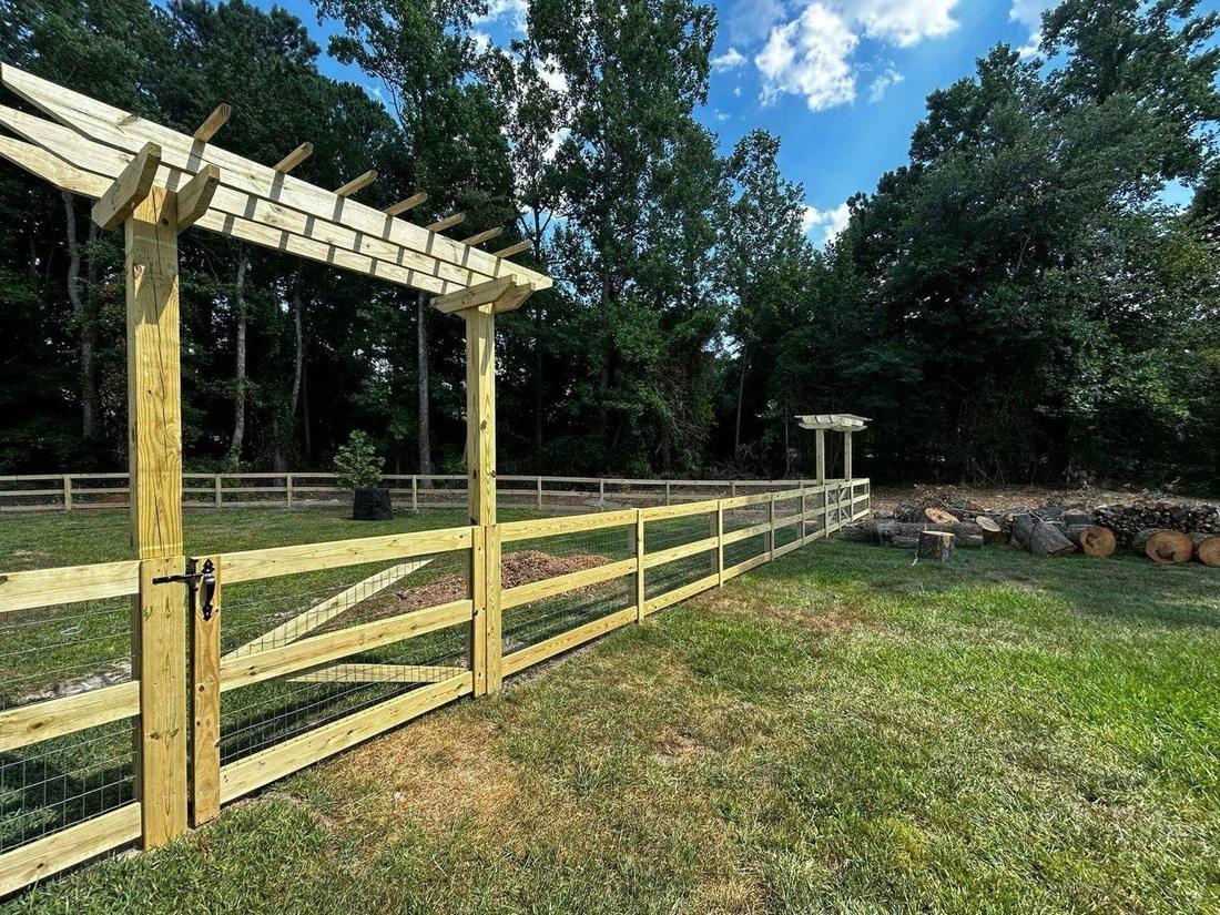 A timber pergola over a gate, joined to a three-rail fence backed with wire mesh.