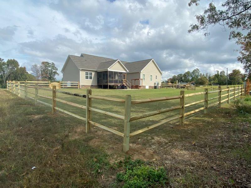 A three-rail wood farm fence with a gate, a house and open pasture behind.