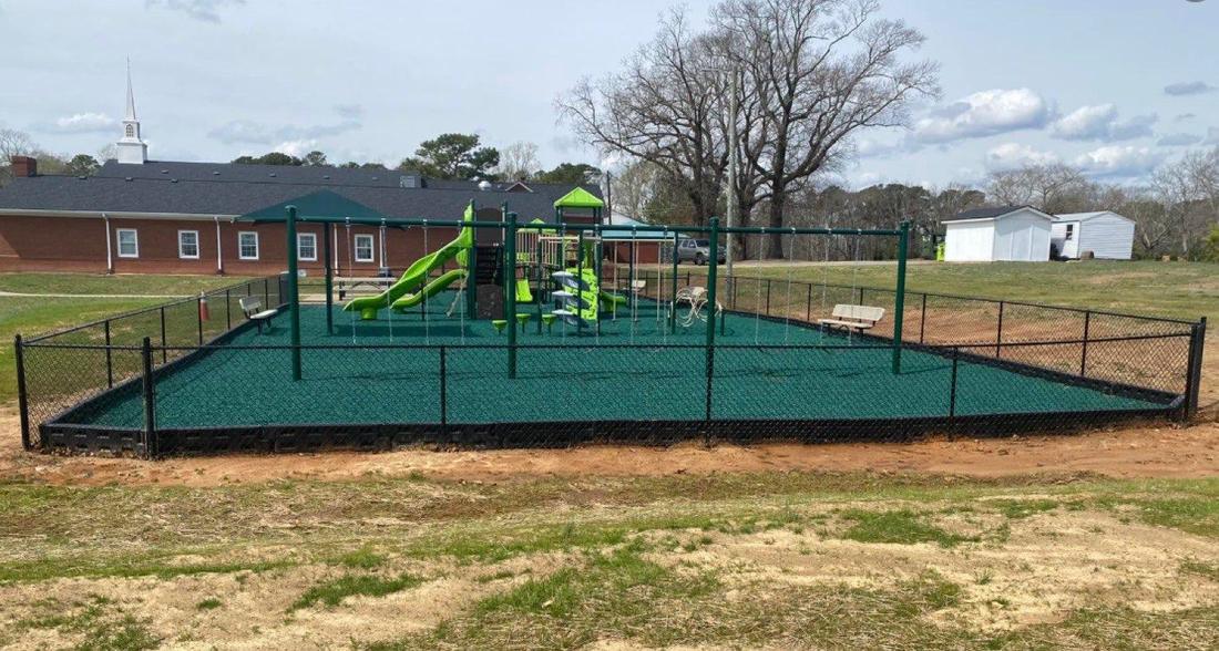 A black fence enclosing a playground with green safety surfacing, a brick building behind.