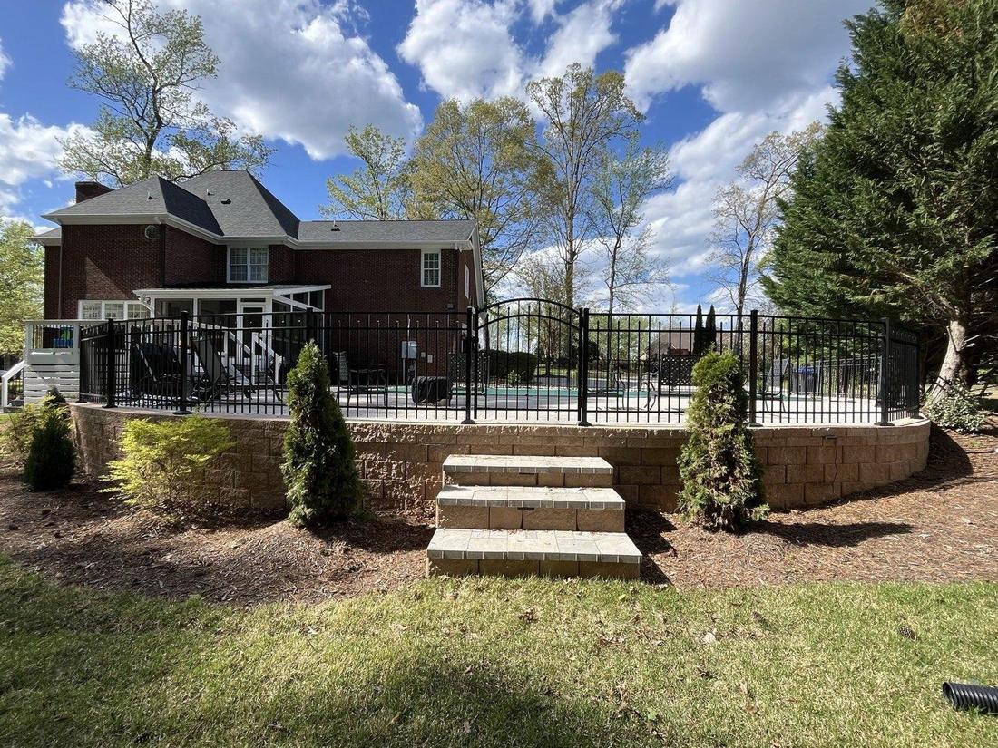A black aluminum fence along the top of a stone retaining wall with steps, a brick house behind.