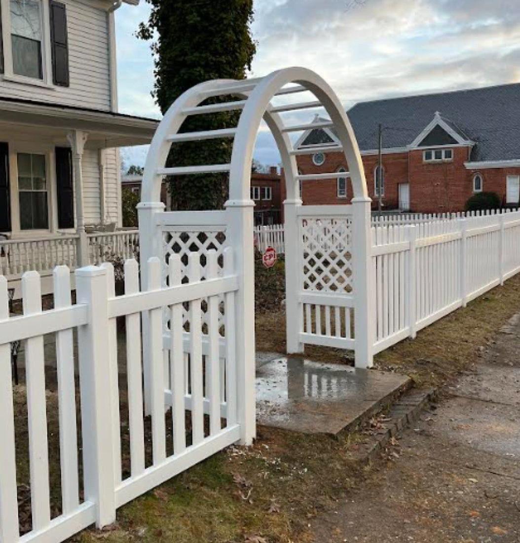 A white vinyl arbor with a lattice arch over a gate, joined to a white picket fence along a path.
