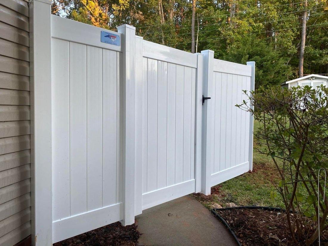 A white vinyl privacy fence and gate running beside a house to a wooded edge.