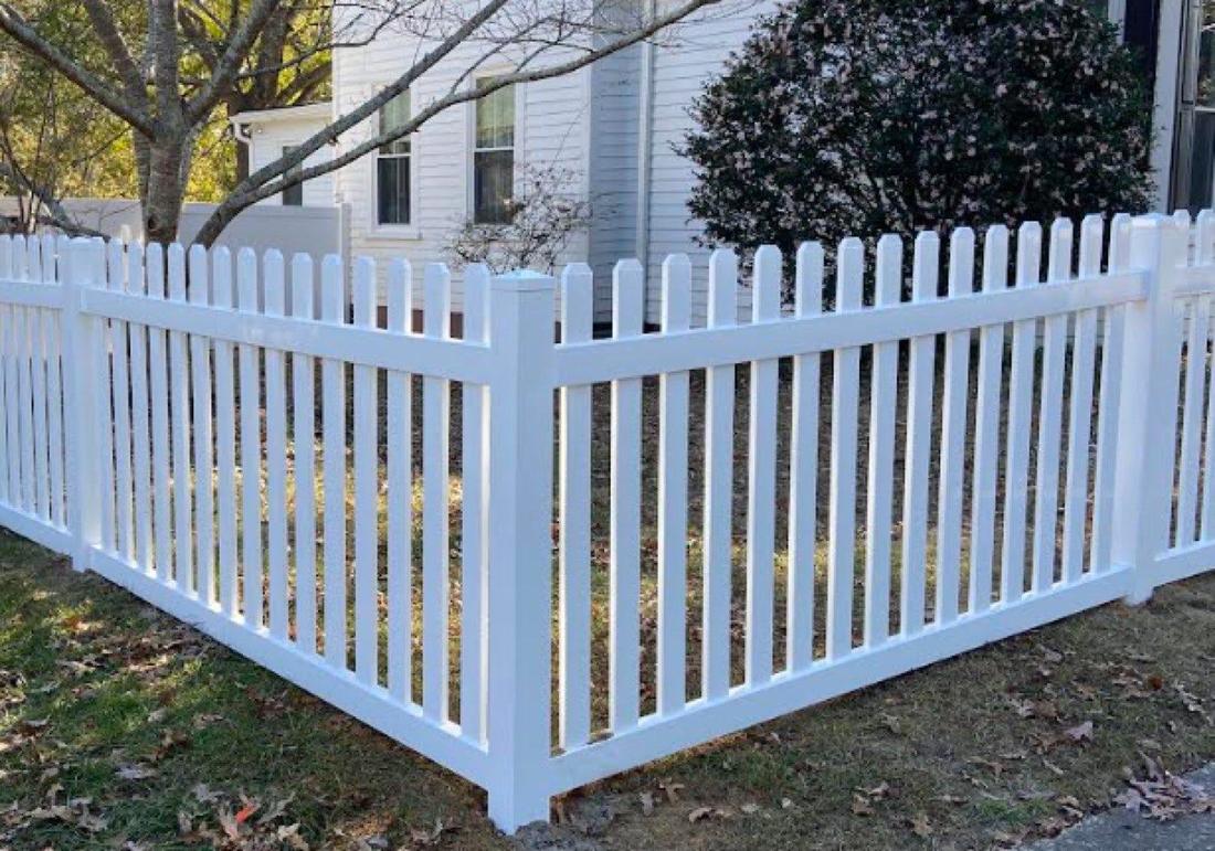 A white vinyl picket fence turning a corner across a lawn, house behind.