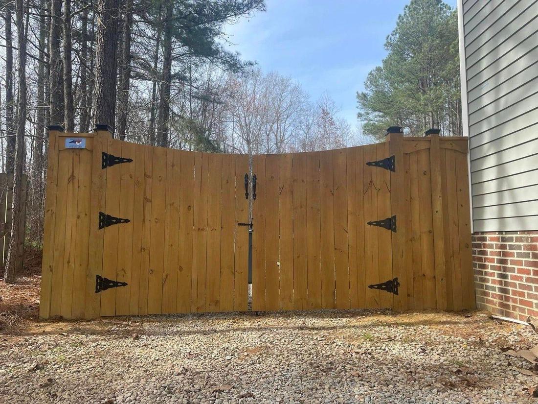 A stained wood double gate with black strap hinges, beside a house on a gravel drive.