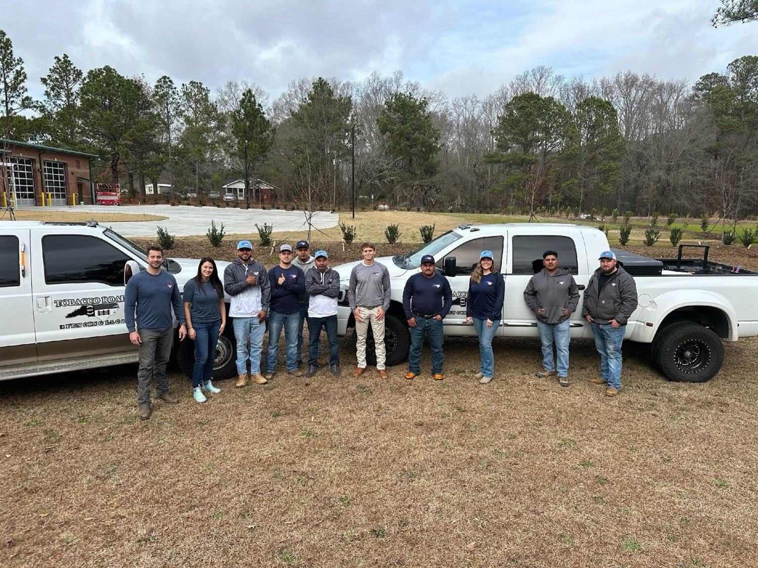 The crew standing in a line in front of two white pickup trucks on bare ground, trees behind.